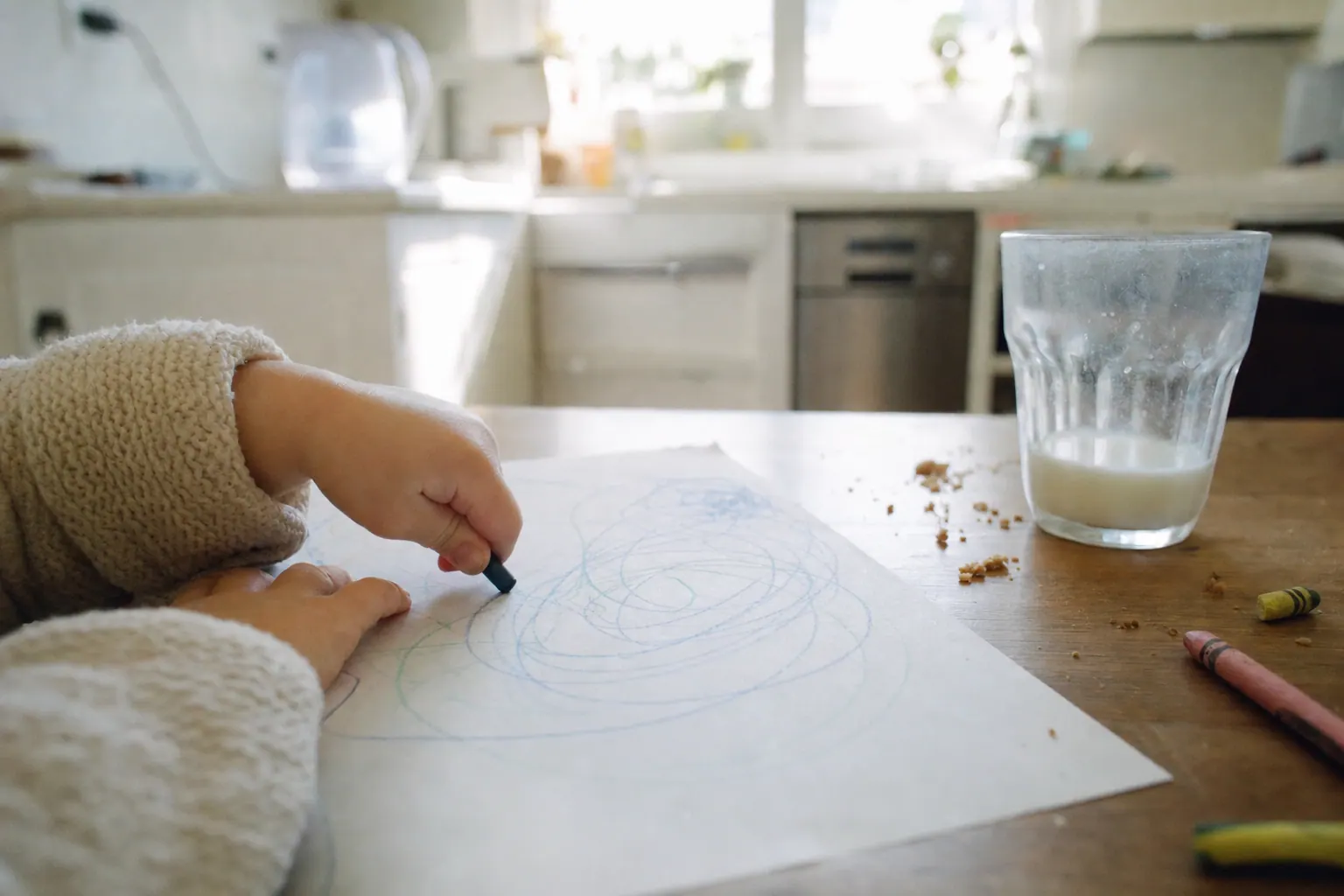 A young child's hands drawing with a crayon on paper at a kitchen table