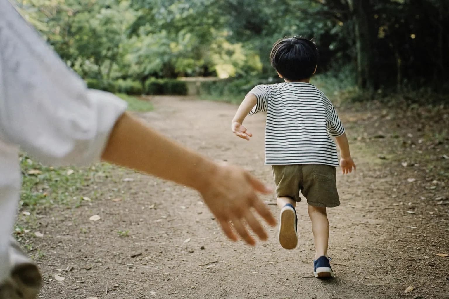 A small child seen from behind, running ahead along a park path as a parent's hand lets go