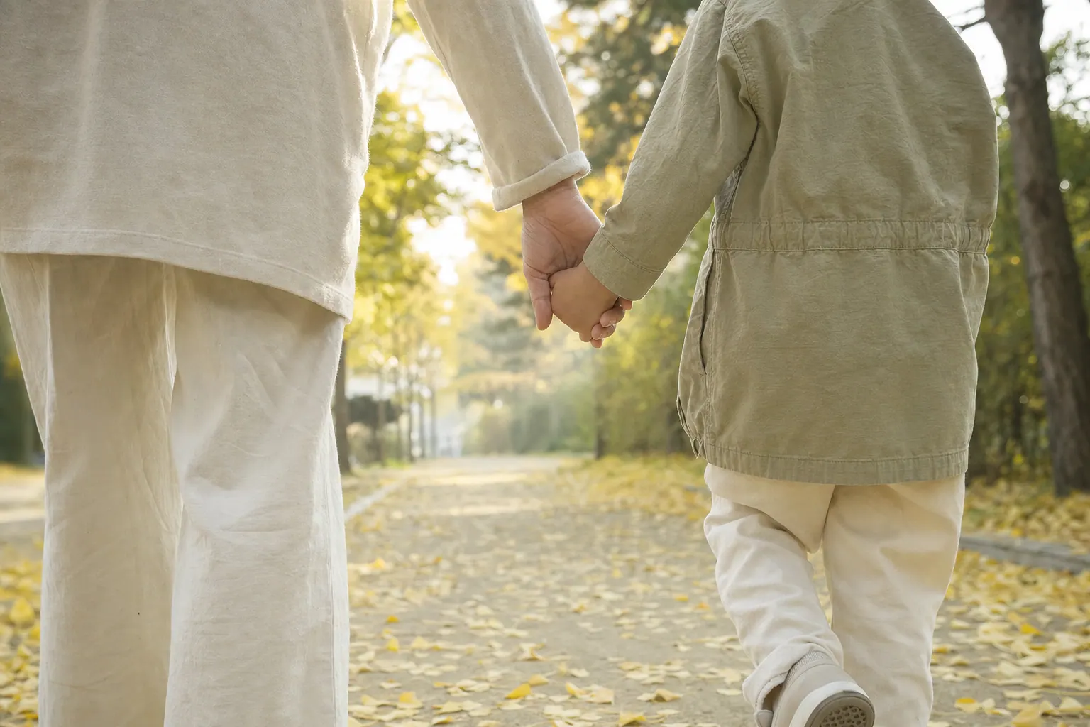 An adult's hand holding a small child's hand as they walk along a tree-lined path