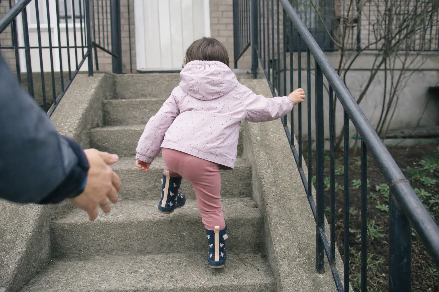 A small child seen from behind, climbing outdoor steps with one hand on the rail and a parent's hand close behind