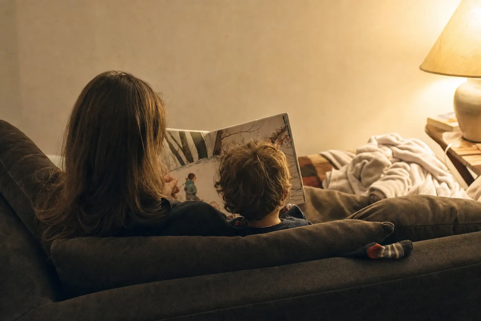A parent and a small child seen from behind on a sofa, reading a picture book together