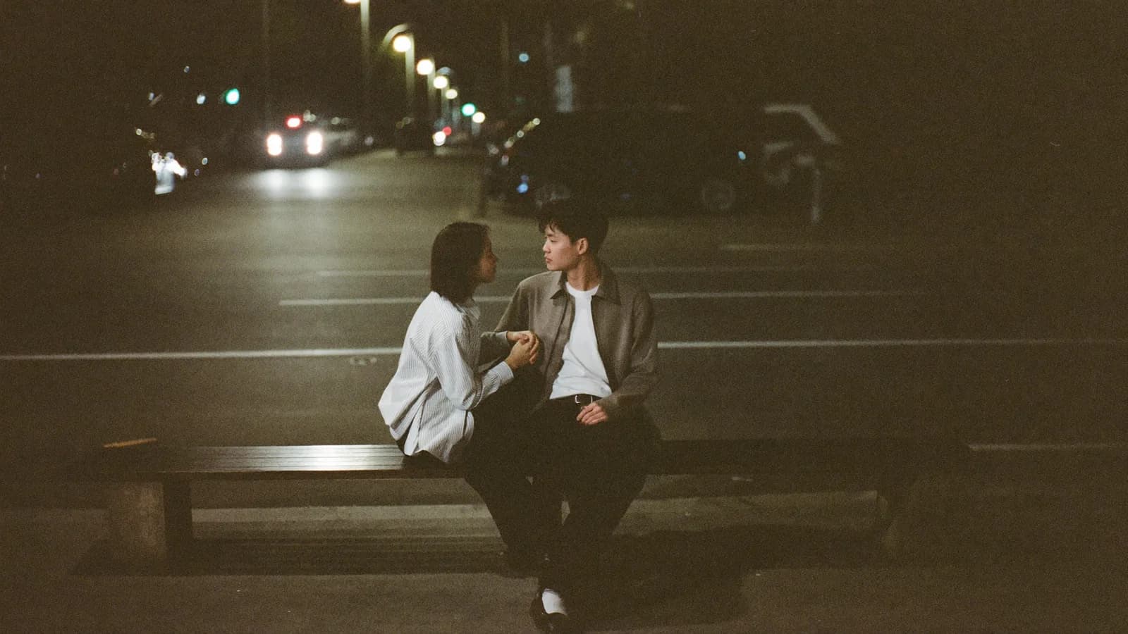 Two people on a night street bench, sitting close, one turned toward the other mid-conversation.